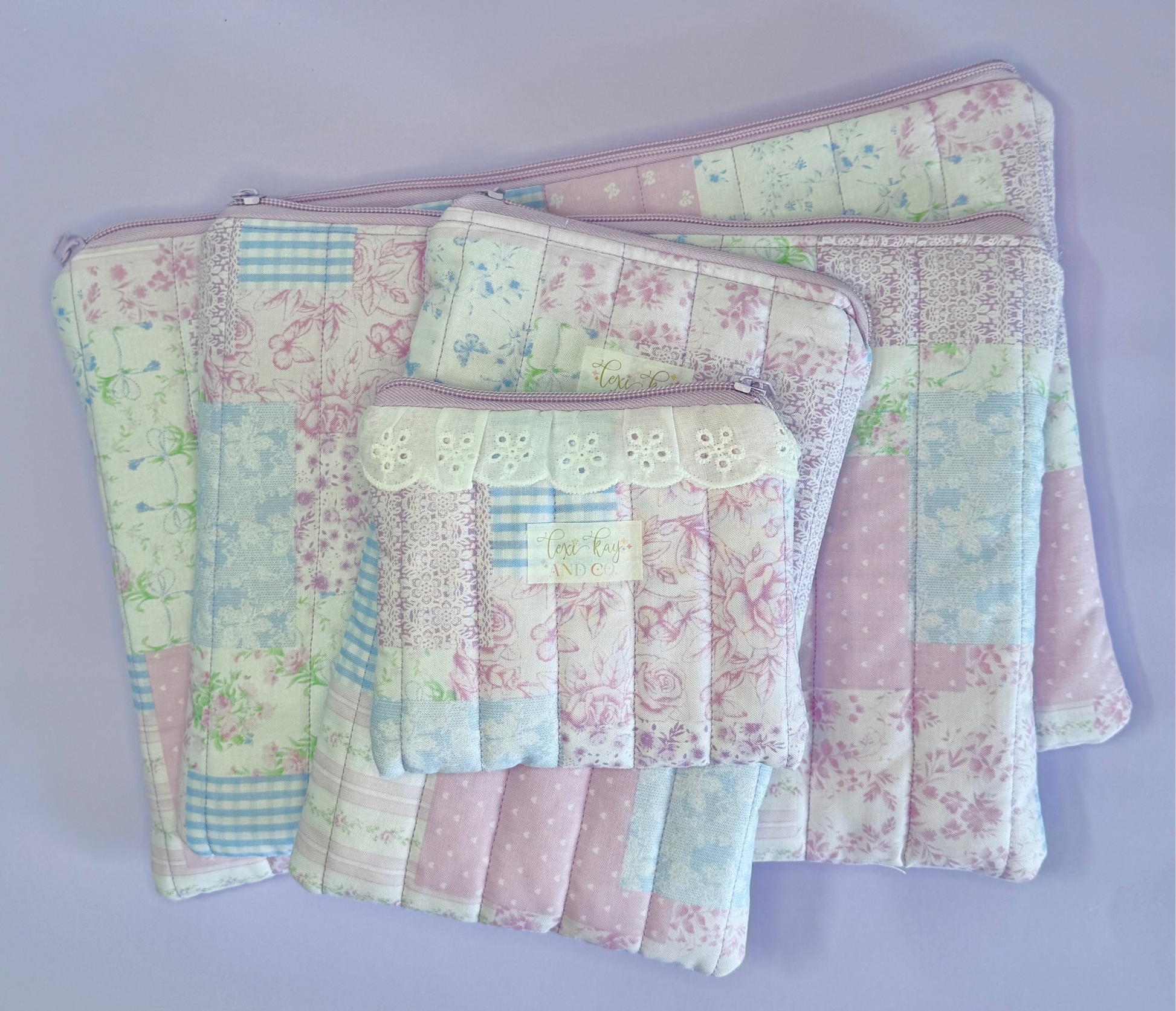 A set of pastel patchwork quilted pouches and small fabric accessories arranged on a light lavender background. The items feature soft pink, blue, and green floral and geometric patterns, with one small pouch decorated with white lace trim.