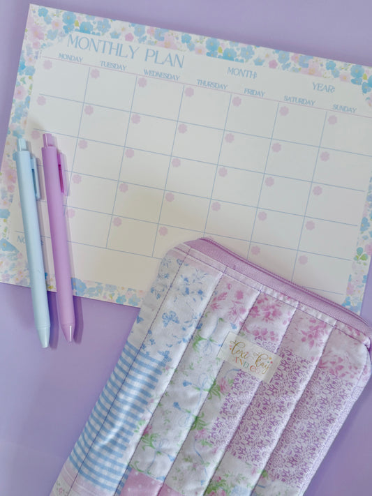 A pastel-themed monthly planner pad with a floral border and dotted calendar grid, styled with two pens and a quilted fabric pouch on a lavender background.