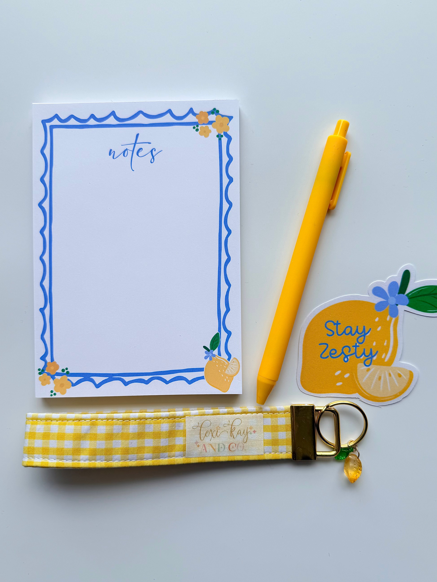A flat-lay of a stationery set on a light gray background, featuring a white notepad with a blue wavy border, a yellow ballpoint pen, a yellow and white gingham fabric wrist strap keychain with gold hardware, and a yellow lemon-shaped sticker that says 'Stay Zesty'.