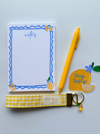 A flat-lay of a stationery set on a light gray background, featuring a white notepad with a blue wavy border, a yellow ballpoint pen, a yellow and white gingham fabric wrist strap keychain with gold hardware, and a yellow lemon-shaped sticker that says 'Stay Zesty'.